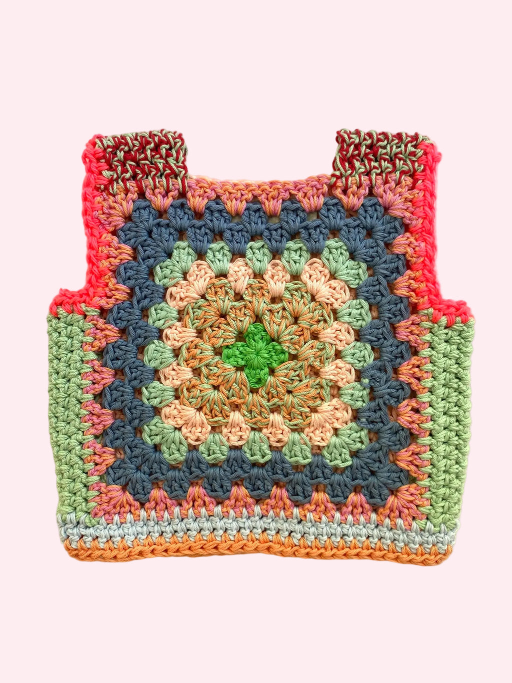 A handmade crocheted granny square vest laid flat on a light beige fabric background. The bag features a multicolor pattern with a central green star, peach and beige concentric circles, a dark blue-gray square, light green borders, neon pink edges, 