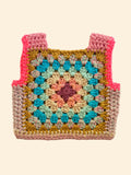A handmade crocheted granny square vest or pouch with pink shoulder straps, featuring a multicolor crochet pattern on a light beige fabric background.