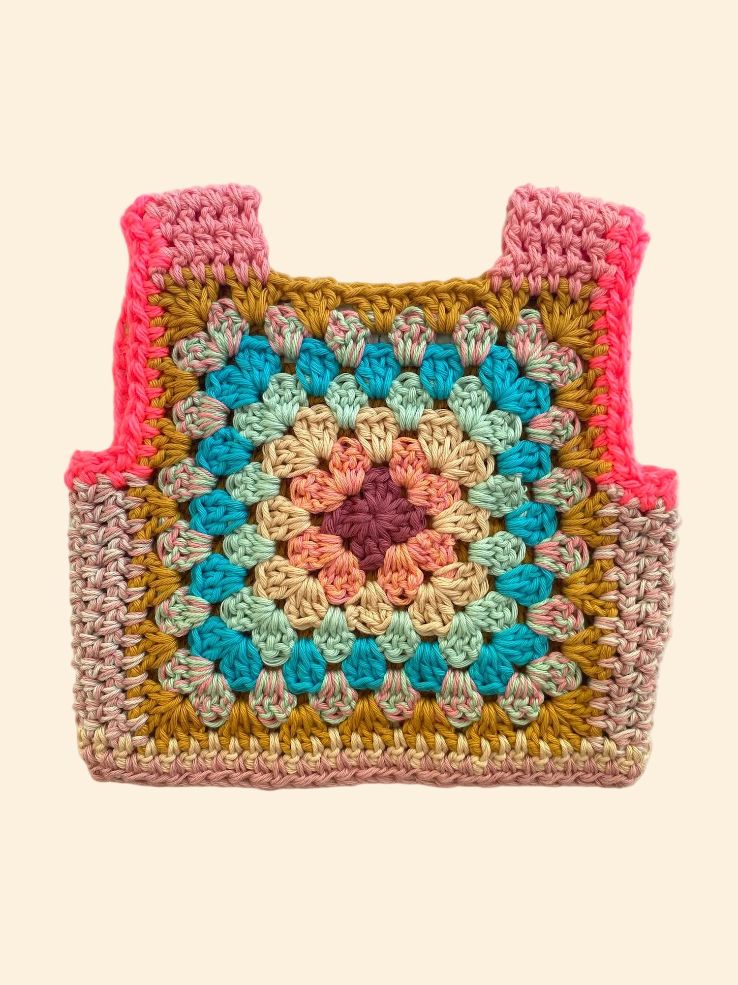 A handmade crocheted granny square vest or pouch with pink shoulder straps, featuring a multicolor crochet pattern on a light beige fabric background.