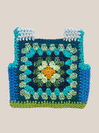 A handmade crocheted baby vest made from a granny square pattern, featuring shades of blue, teal, green, and a yellow-orange center, laid flat on a light beige textured background.