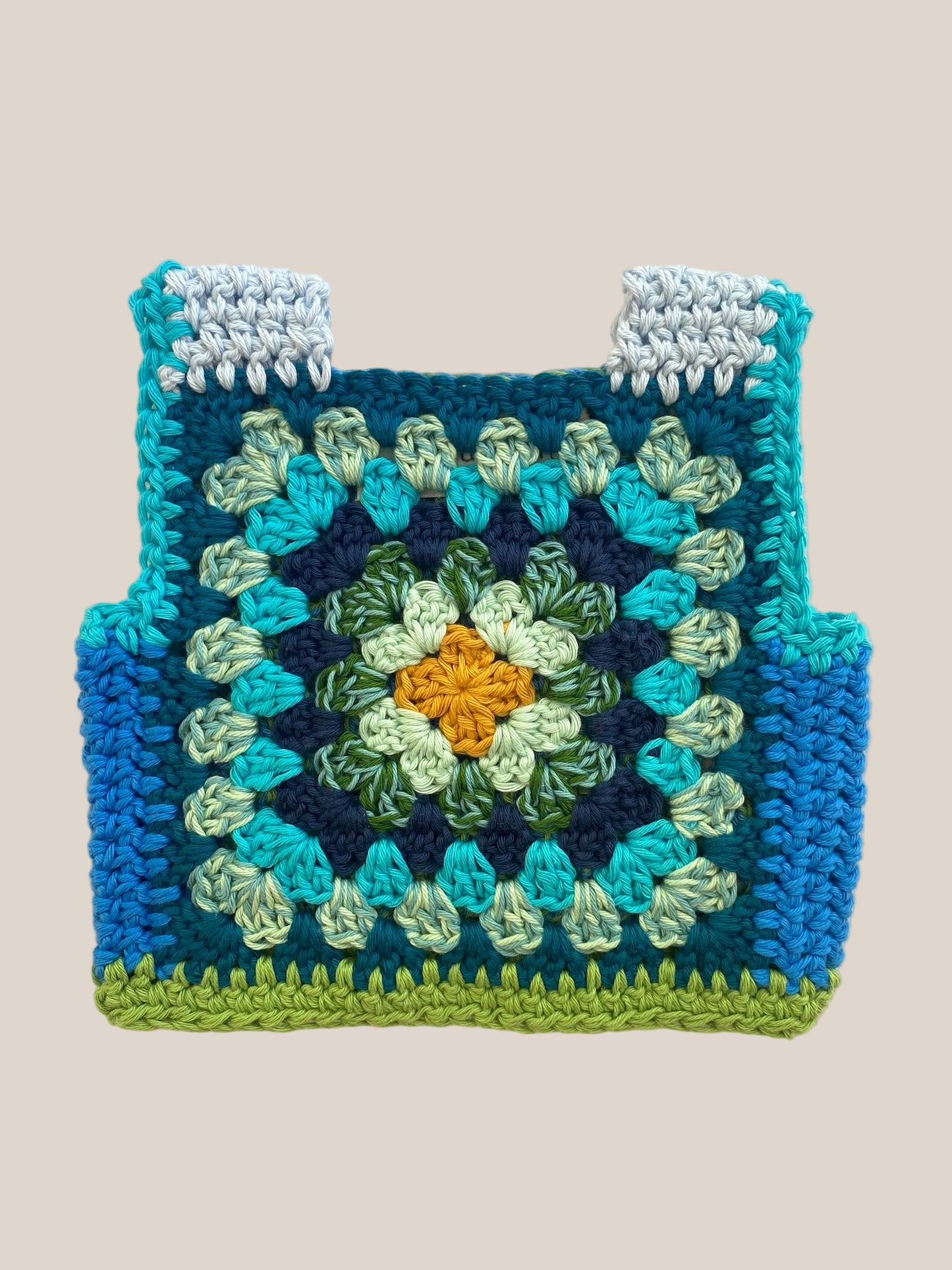 A handmade crocheted baby vest made from a granny square pattern, featuring shades of blue, teal, green, and a yellow-orange center, laid flat on a light beige textured background.