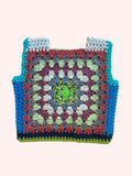 A handmade crocheted square vest  laid flat on a light beige fabric background. The item features a multicolor granny square pattern with concentric rings of green, light blue, purple, gray, and rust red, finished with a solid turquoise blue border.