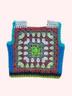 A handmade crocheted square vest  laid flat on a light beige fabric background. The item features a multicolor granny square pattern with concentric rings of green, light blue, purple, gray, and rust red, finished with a solid turquoise blue border.