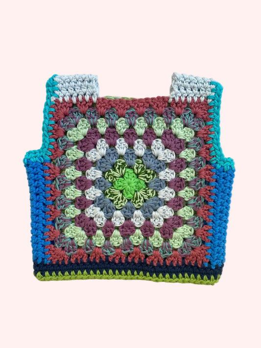 A handmade crocheted square vest  laid flat on a light beige fabric background. The item features a multicolor granny square pattern with concentric rings of green, light blue, purple, gray, and rust red, finished with a solid turquoise blue border.