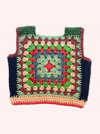 A handmade crocheted granny square vest or top laid flat on a light beige fabric background. The garment features a multicolor pattern with green, red, blue, pink, and brown yarn, finished with dark navy blue side panels and a peach-colored bottom trim.