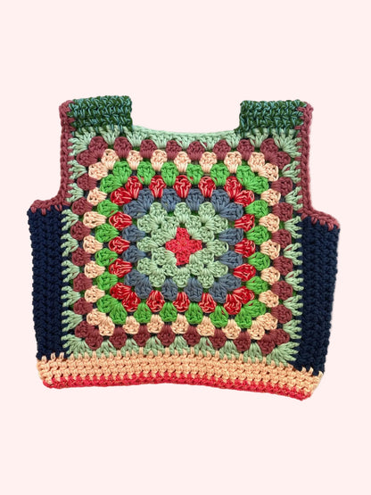 A handmade crocheted granny square vest or top laid flat on a light beige fabric background. The garment features a multicolor pattern with green, red, blue, pink, and brown yarn, finished with dark navy blue side panels and a peach-colored bottom trim.
