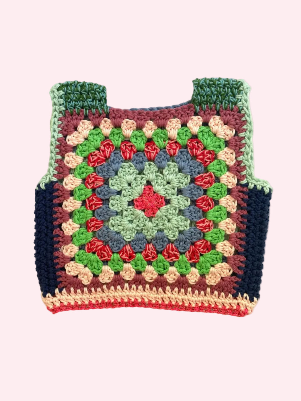 A handmade crocheted granny square vest laid flat on a light beige fabric background. The vest features a multicolor pattern with green, blue, red, pink, and dark navy blue yarn, complete with shoulder straps.