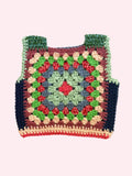 A handmade crocheted granny square vest laid flat on a light beige fabric background. The vest features a multicolor pattern with green, blue, red, pink, and dark navy blue yarn, complete with shoulder straps.
