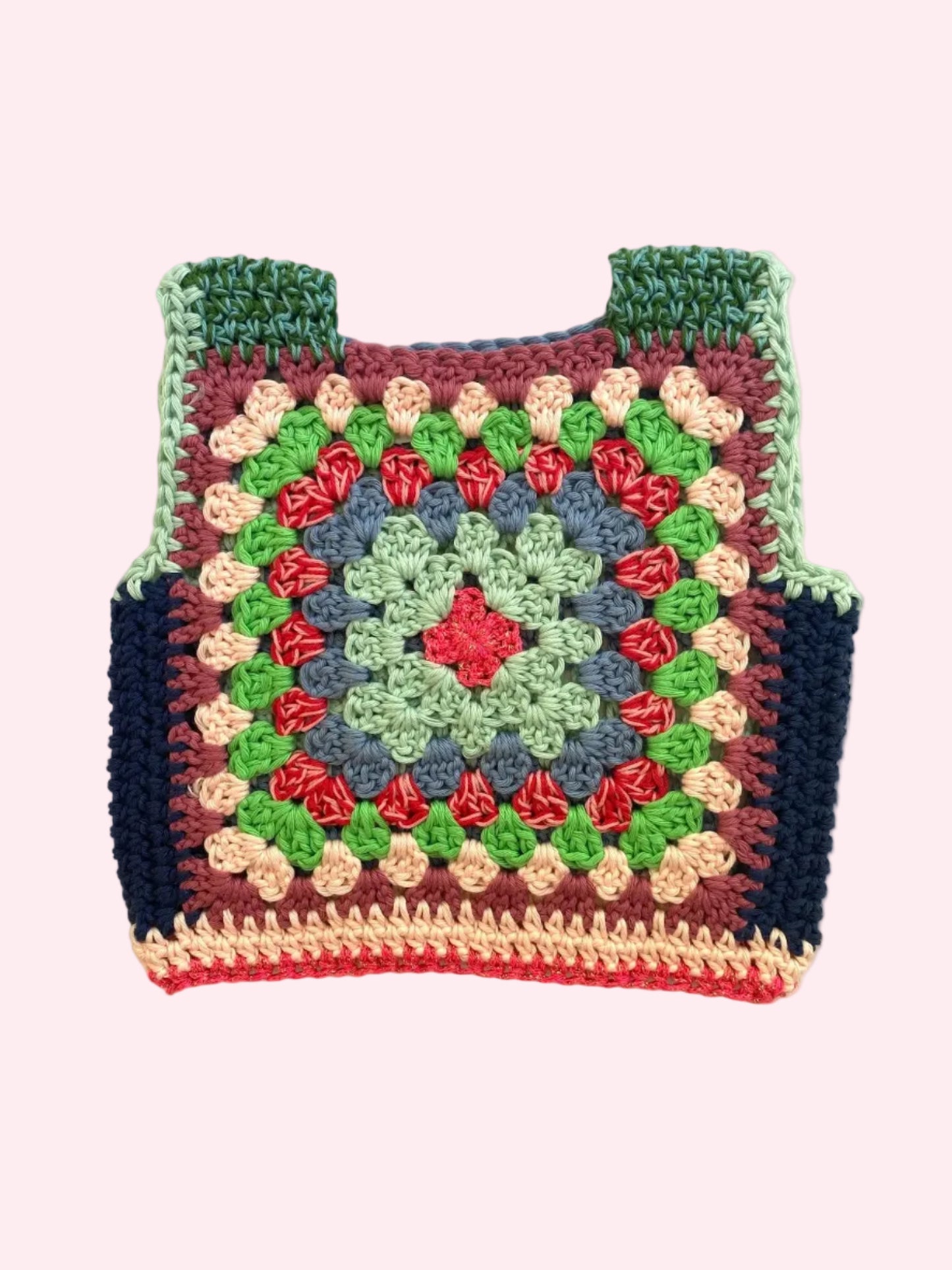A handmade crocheted granny square vest laid flat on a light beige fabric background. The vest features a multicolor pattern with green, blue, red, pink, and dark navy blue yarn, complete with shoulder straps.