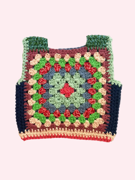 A handmade crocheted granny square vest laid flat on a light beige fabric background. The vest features a multicolor pattern with green, blue, red, pink, and dark navy blue yarn, complete with shoulder straps.