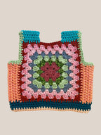 A handmade crocheted granny square vest laid flat on a light beige fabric background. The bag features a multicolor pattern with a central pink and green medallion, surrounded by blue, brown, and peach sections.
