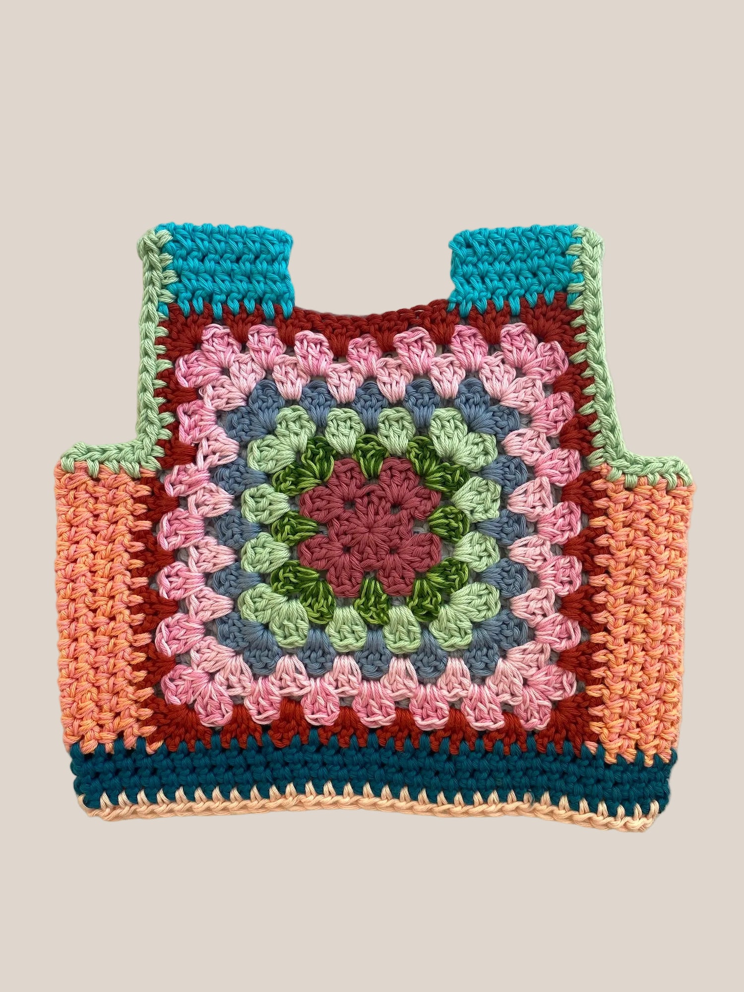 A handmade crocheted granny square vest laid flat on a light beige fabric background. The bag features a multicolor pattern with a central pink and green medallion, surrounded by blue, brown, and peach sections.