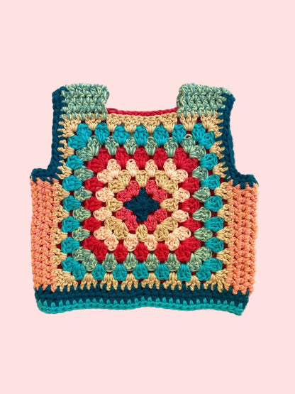 A handmade crocheted vest or top with a granny square pattern, featuring teal, red, beige, and peach colors, laid flat on a light beige fabric background.