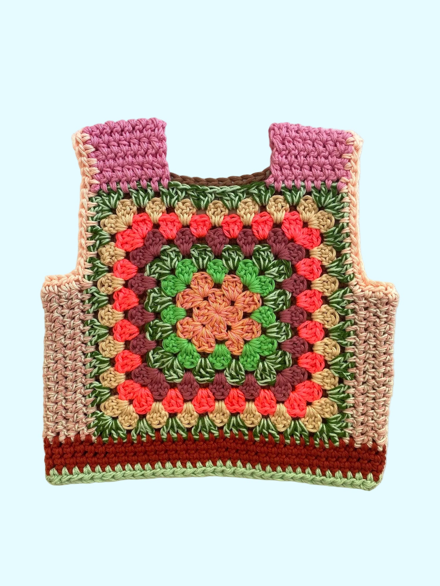 A handmade crocheted vest with a colorful granny square pattern, featuring pink shoulder panels, green and pink floral motifs, and a brown base, laid flat on a light beige fabric background.