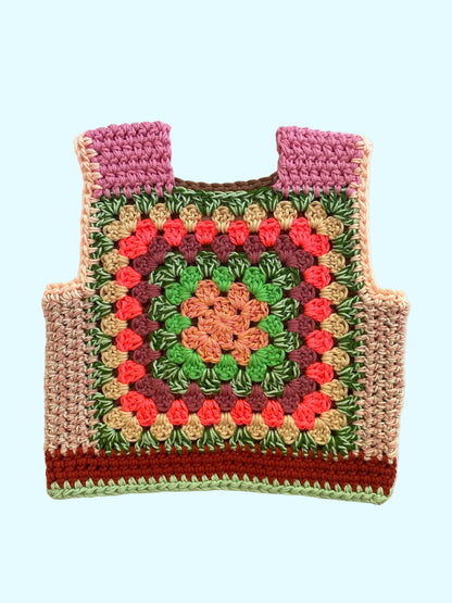 A handmade crocheted vest with a colorful granny square pattern, featuring pink shoulder panels, green and pink floral motifs, and a brown base, laid flat on a light beige fabric background.